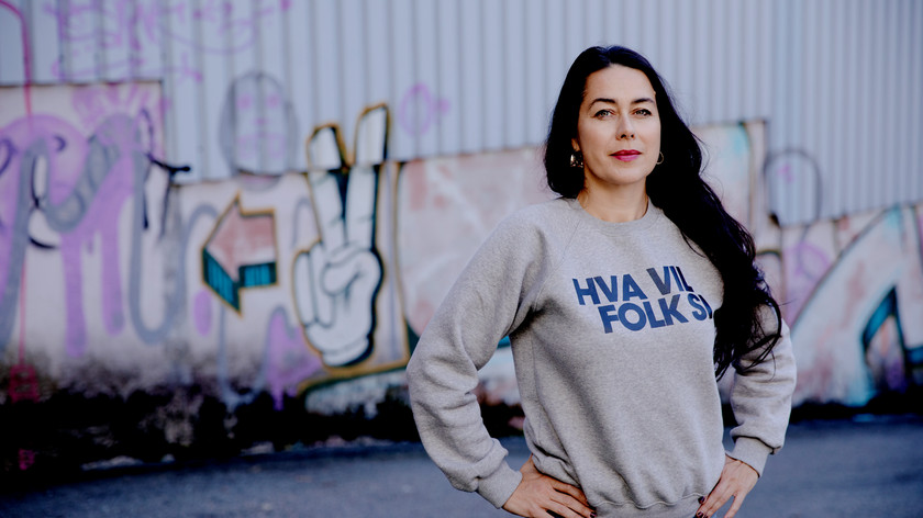 Foto. En kvinne med langt mørkt hår ser mot kamera. Hun har på seg en genser med påskriften Hva vil folk si. Bak henne en vegg med grafitti.