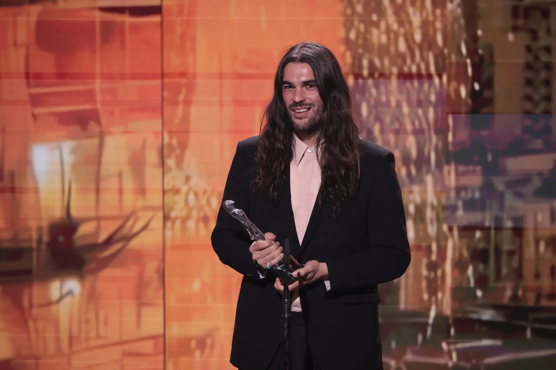 Foto. En langhåret mann i dress holder en statuett på en scene.