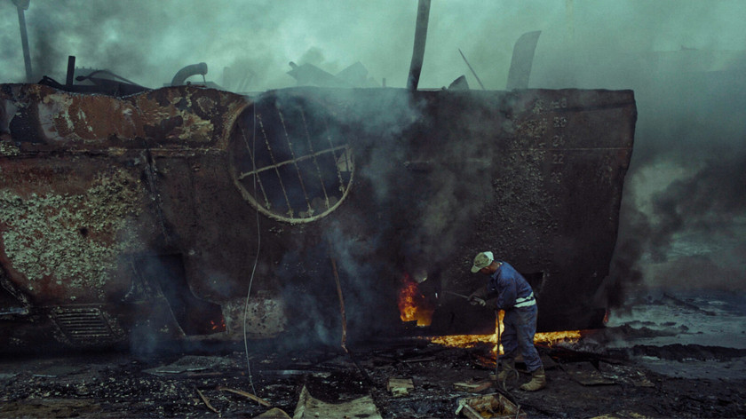Foto. Foran et brennende skipsvrak står en mann og jobber.
