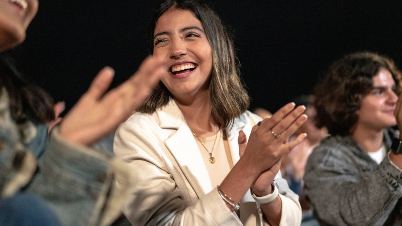 Foto. Flere personer i en sal klapper. I fokus en ung dame i hvit dressjakke.