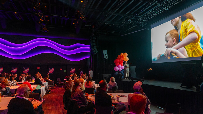 Foto. Grupper med mennesker rundt store runde bord i en sal ser på et stort lerret.
