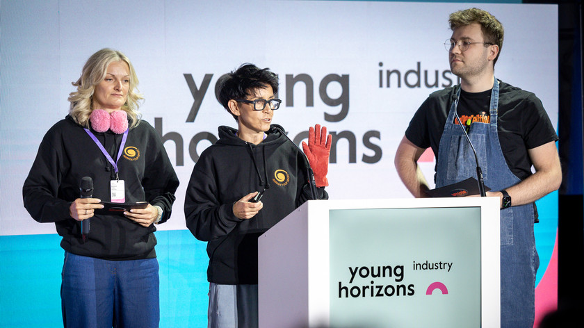 Foto. Tre personer, to damer og en mann, står ved en talerstol.