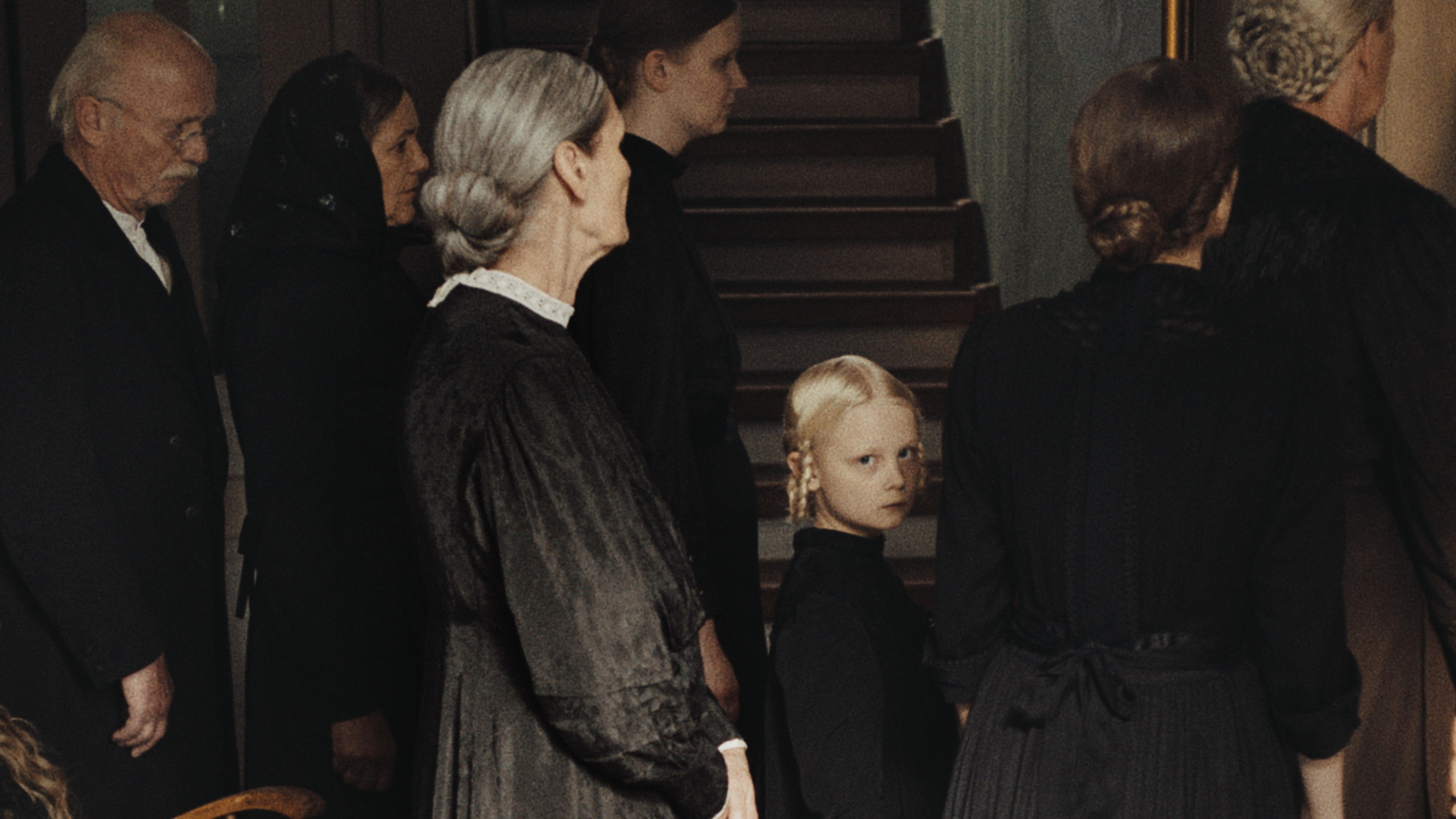 Foto. En gruppe personer kledd i sort sett fra siden eller med ryggen til. En liten jente med fletter vender seg mot kamera. 