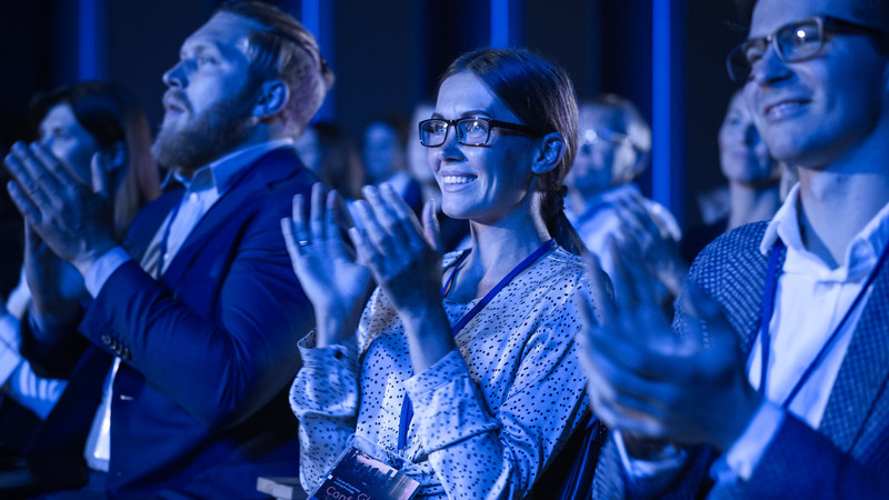 Foto. En forsamling personer i en sal som klapper. I fokus en dame med briller som sitter mellom to menn i dress. 