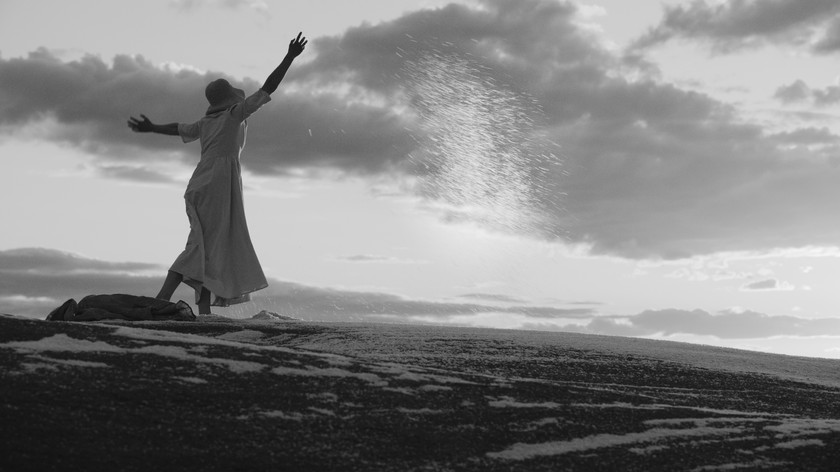 Foto. Sort/hvitt. En dame i lang kjole og hatt står med ryggen til på en haug med armene utslått. Over henne er det himmel med skyer.