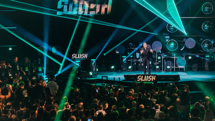 Foto. En mann står på scenen foran prublikum. Stråler med farget lys i ulike retninger fra scenen. Slush-logoen vises flere steder.