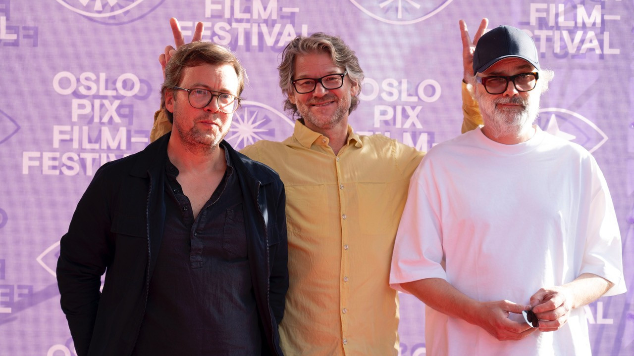 Foto. Tre menn med briller foran en fotovegg. 