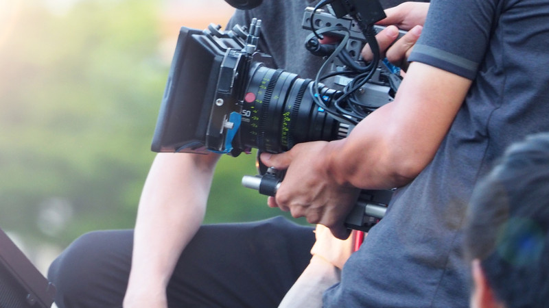 Foto. Et filmkamera holdes av en mann. Rundt skimtes flere personer.