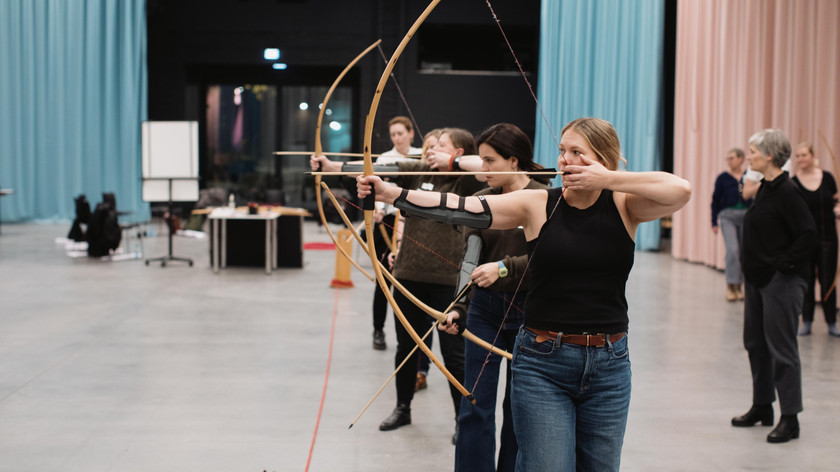 Foto. Flere damer med pil og bue på en rekke i et stort rom. 