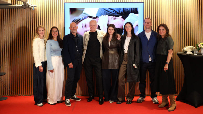 Foto. Åtte personer står på en rekke på rød løper foran en stor skjerm.