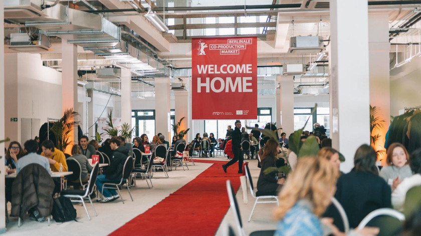 Foto. Gruppemøter rundt flere bord i et stort lyst rom. En banner henger fra taket over en rød løper på gulvet.