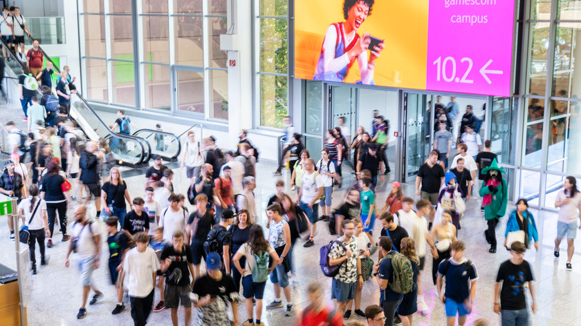 Foto. En stor gruppe med mennesker beveger seg rundt i et bygg med glassvegger og rulletrapp. En stor skjerm lyser viser vei.