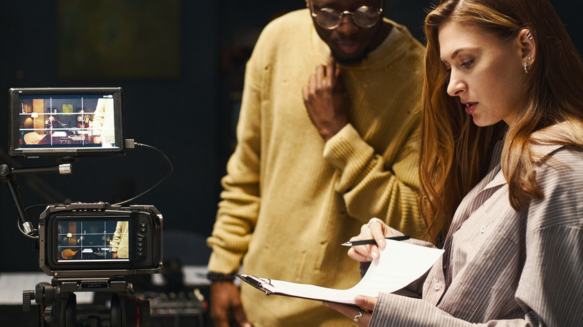Foto. En mann og en dame står ved siden av et filmkamera som filmer. Hun har en blokk og en penn som de begge ser på.
