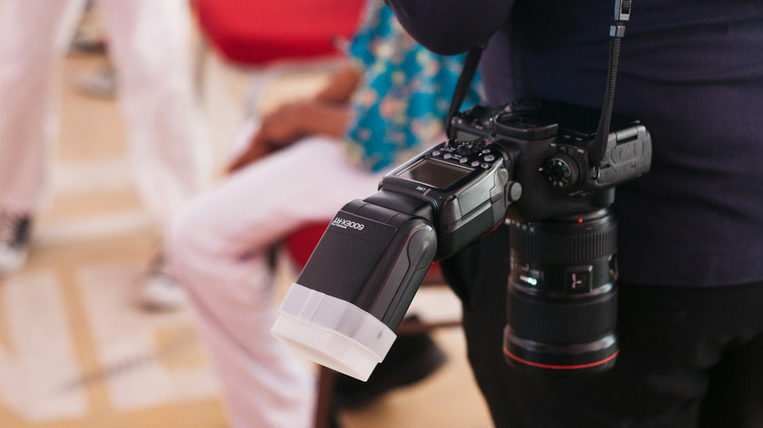 Foto. Et fotoapparat som henger over skulderen på en person. I bakgrunnen skimtes to personer i hvite bukser.