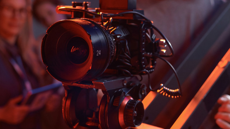 Foto. Filmkamera sett skrått fra siden. Personer skimtes i bakgrunnen