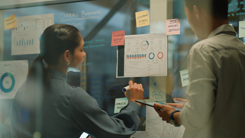 Foto. To personer står med ryggen til og skriver på en tavle. På tavla henger flere lapper og ark. 