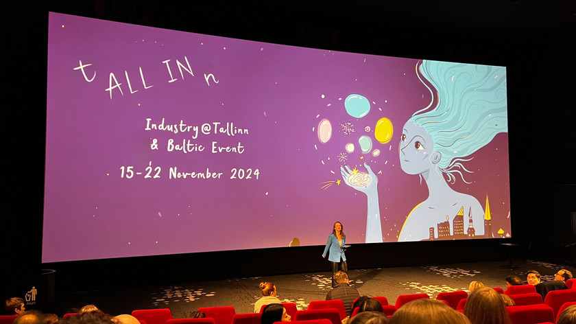 Foto. En dame står på scenen i en kinosal med røde seter. Bak henne er et lerret med artworket til festivalen.