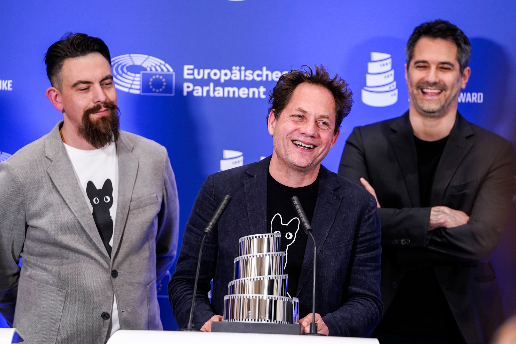 Foto. Tre menn smiler, den ene holder en pris i hendene.