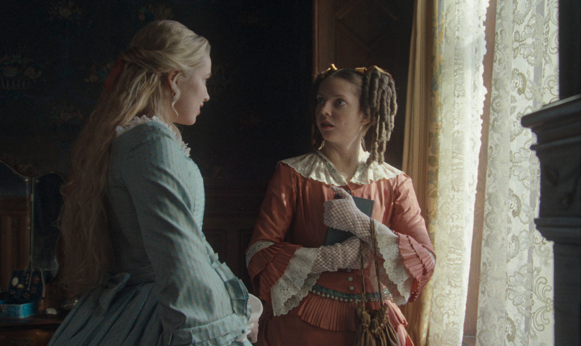 Foto. To unge kvinner i epoke-riktige kjoler står ved et vindu og snakker sammen. 