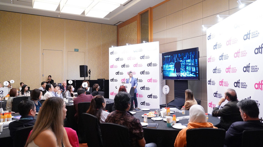 En gruppe mennesker ved runde bord følger med på en mann som presenterer et prosjekt. Bak mannen er en Asia TV Forum & Market-logovegg og en TV. Foto.