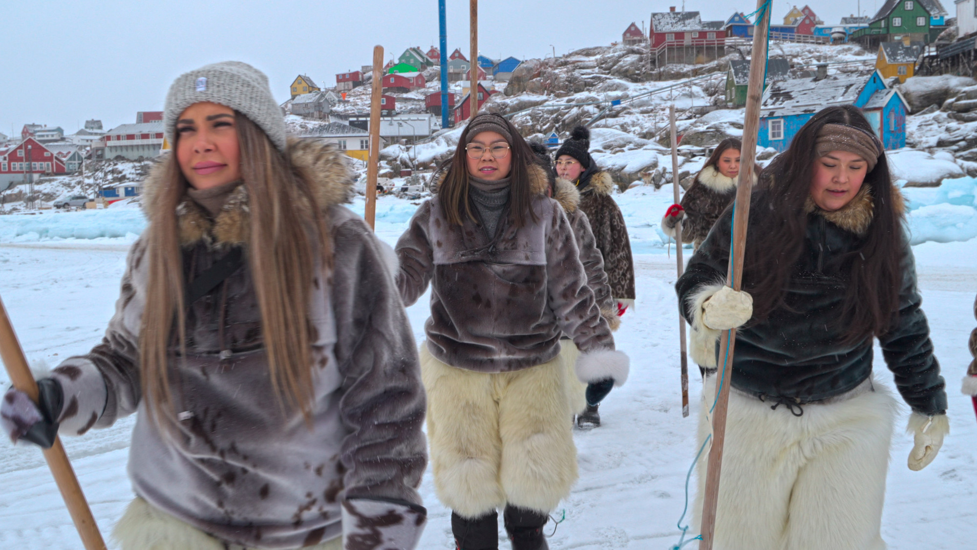 Foto. En gruppe kvinner i selskinnsjakker går med en stav i hånda. I bakgrunnen tykk is og bebyggelse med fargerike hus på en liten topp.