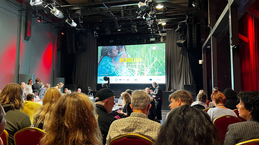 Foto. En gruppe mennesker er samlet i et stort lokale. De sitter på stoler foran et stort lerret.