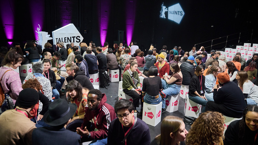 Foto. En stor gruppe unge mennesker sitter på små pappkrakker og snakker sammen i mindre grupper.