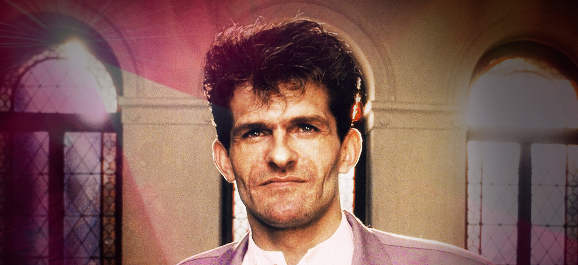 Foto. Portrett av en mann med mørkt hår. Buende vinduer i bakgrunnen som slipper inn lys.