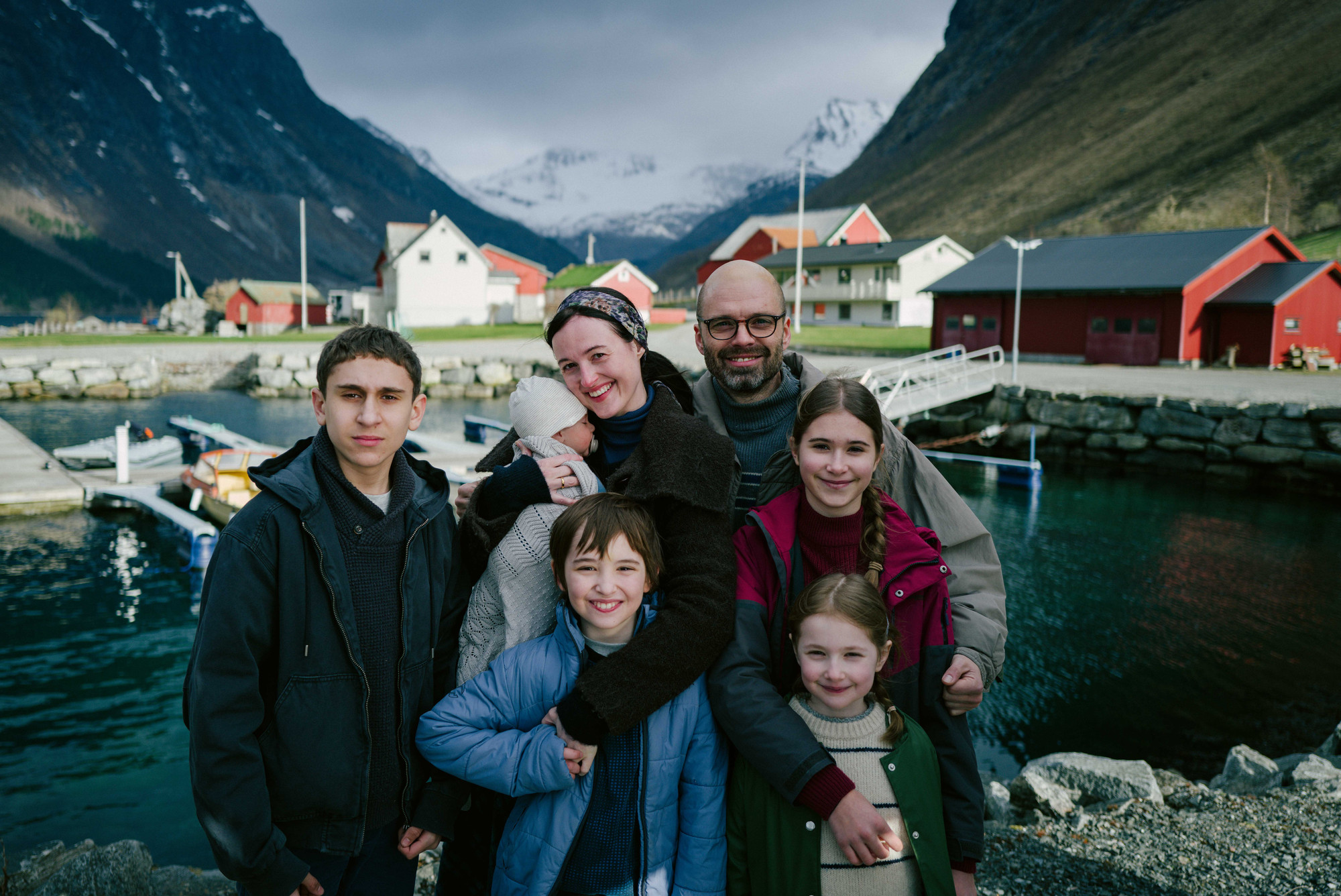 Foto. En familie med to voksne og fem barn står ved et vann med fjell i bakgrunnen. 