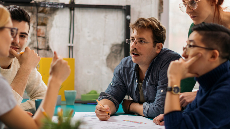 Foto. En gruppe mennesker sitter rundt et bord og prater. På bordet ligger div. papirer. En mann holder en penn.