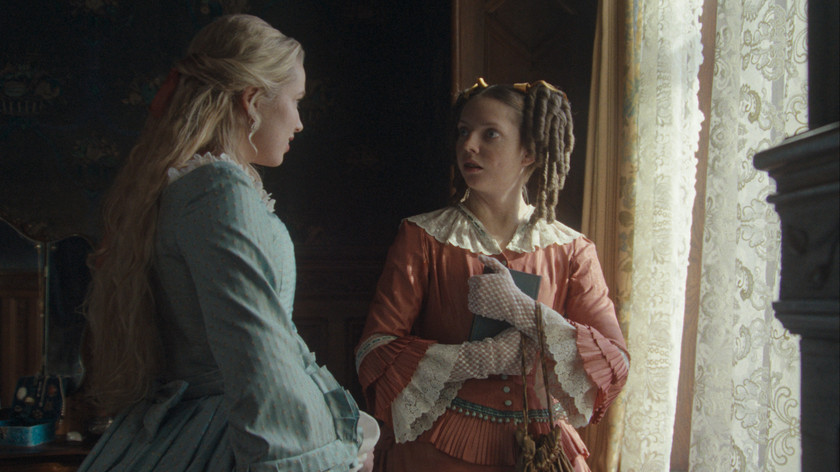 Foto. To unge kvinner i epoke-riktige kjoler står ved et vindu og snakker sammen. 