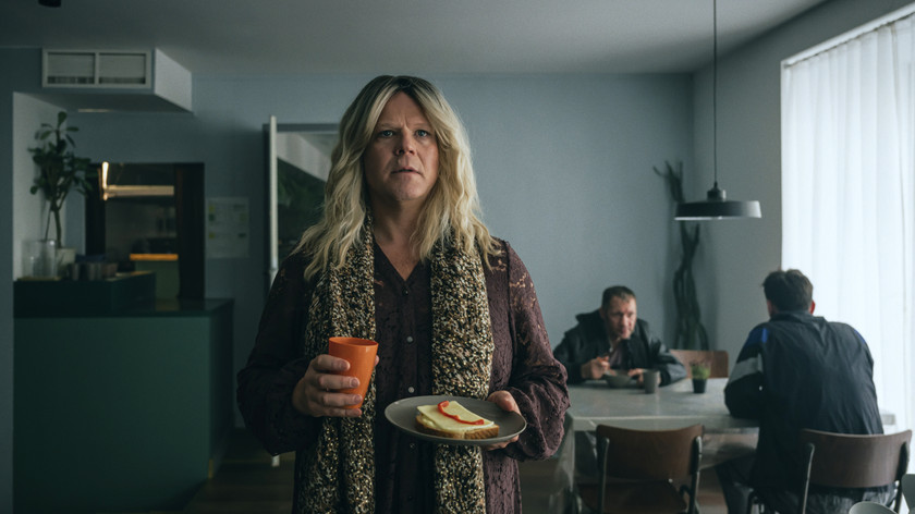 Foto. En mann i kvinneklær holder en asjett med ei brødskive og en kopp. I bakgrunnen sitter to menn ved et bord.