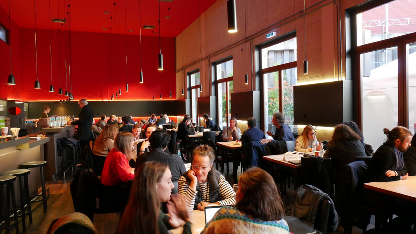 Foto. Små grupper på tre personer sitter rundt mange bord i en kafe. En lang bardisk vises til venstre. En vindusrekke til høyre.