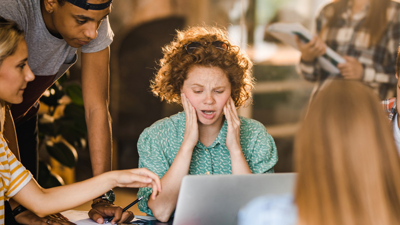 Foto. En dame med krøllete hår ser oppgitt ned på en datamaskin, rundt henne sitter og står det en gruppe personer.