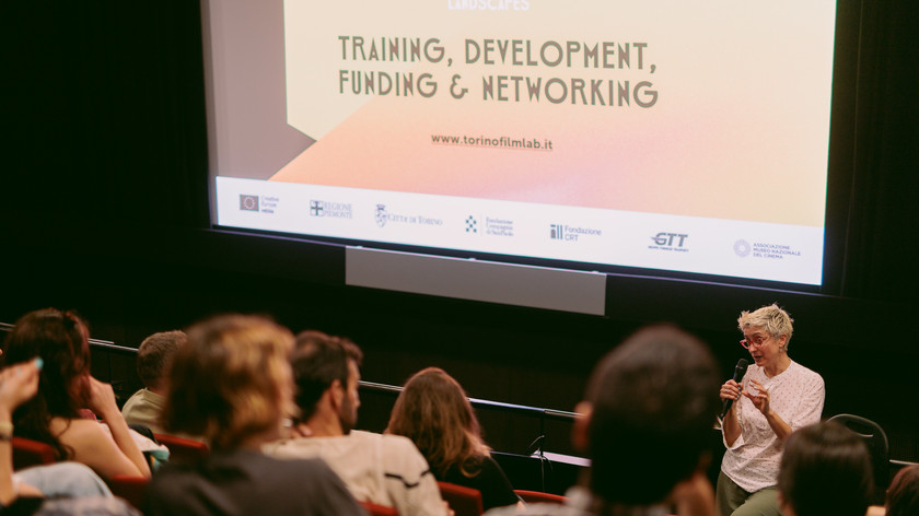 Foto. Publikum i en sal. Ei dame snakker i en mikrofon foran et stort lerret.