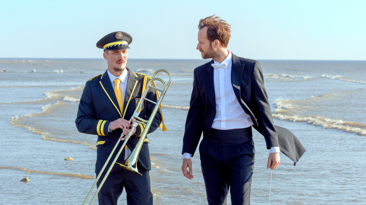Foto. To menn, en i korpsuniform og en i sjakett, går i vannkanten. Den ene holder en trombone.