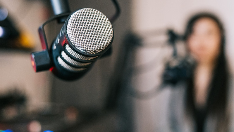 Foto. En mikrofon i et studio. I bakgrunnen skimtes en dame bak en annen mik.